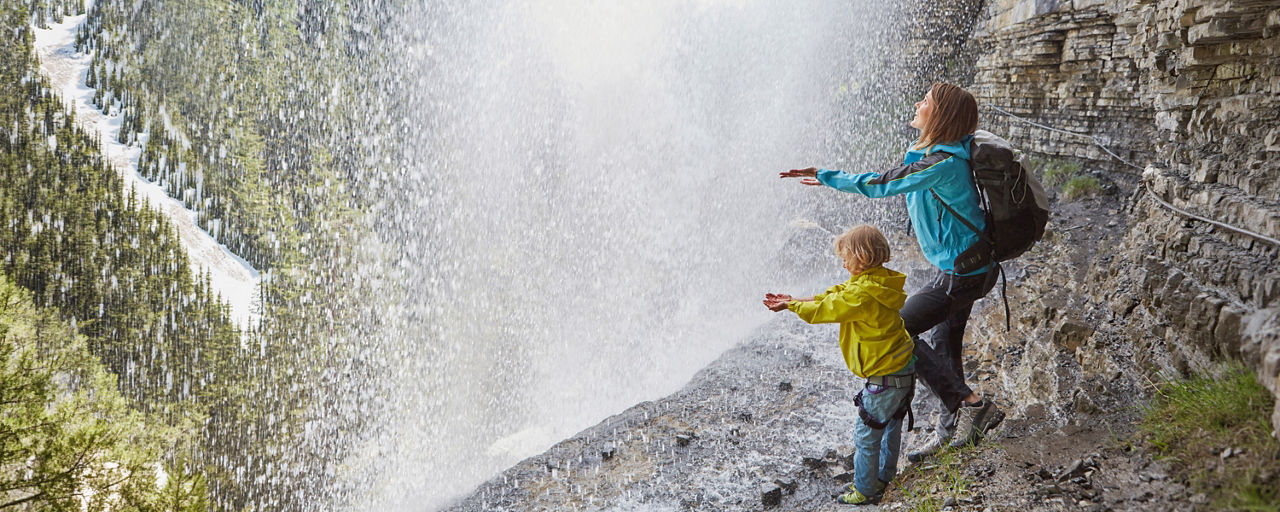 Mutter und Kind stehen mit ausgestreckten Händen im Gebirge unter einem Wasserfall