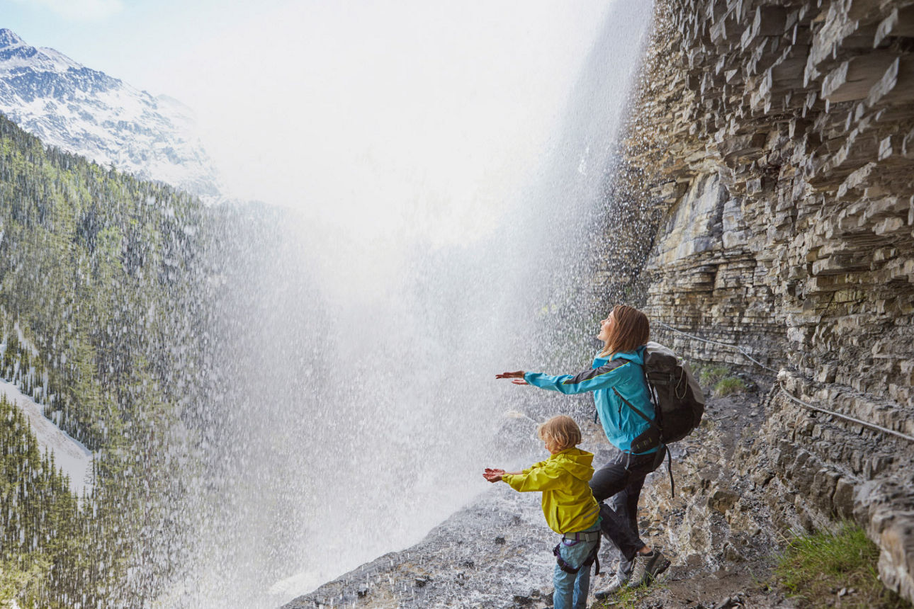 Mutter und Kind stehen mit ausgestreckten Händen im Gebirge unter einem Wasserfall
