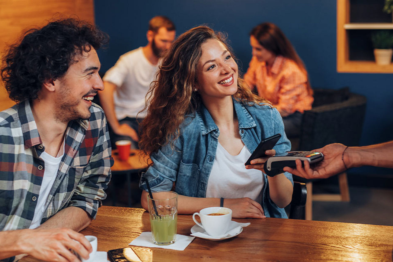 Zwei junge Menschen lachen und bezahlen kontaktlos mit Karte im Café