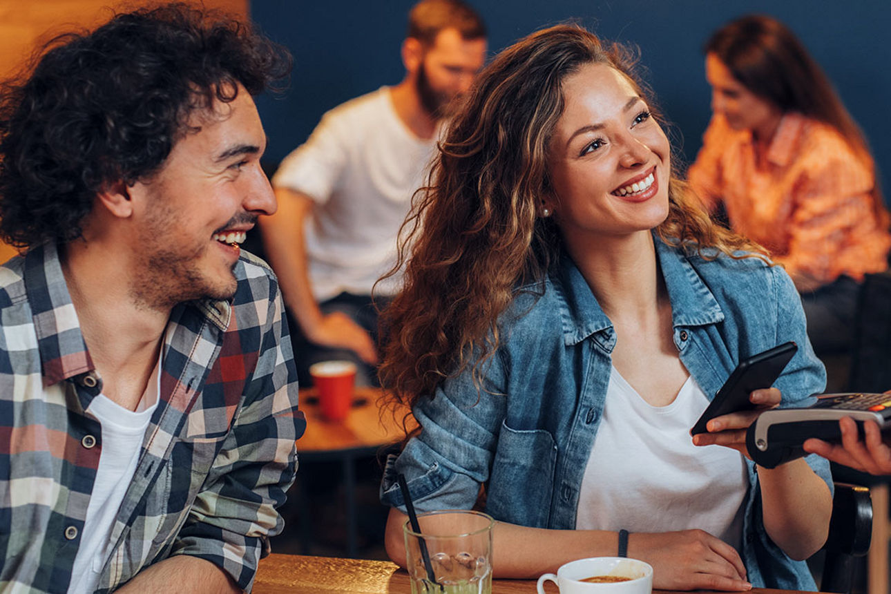 Zwei junge Menschen lachen und bezahlen kontaktlos mit Karte im Café