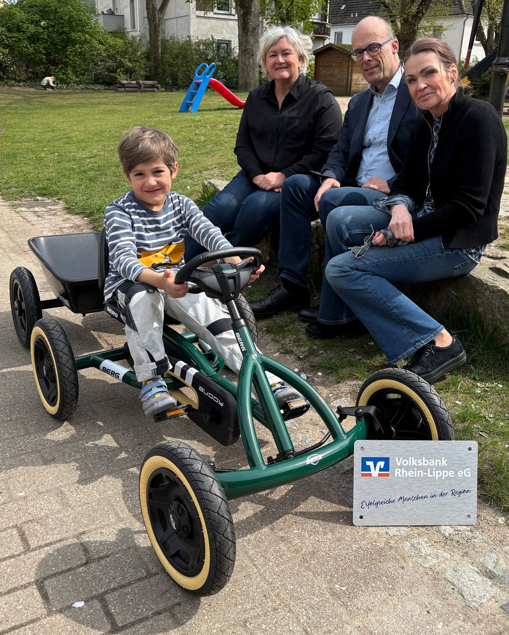 Ein Junge fährt fröhlich auf einem grünen Kettcar, während drei Erwachsene lächelnd im Hintergrund sitzen.