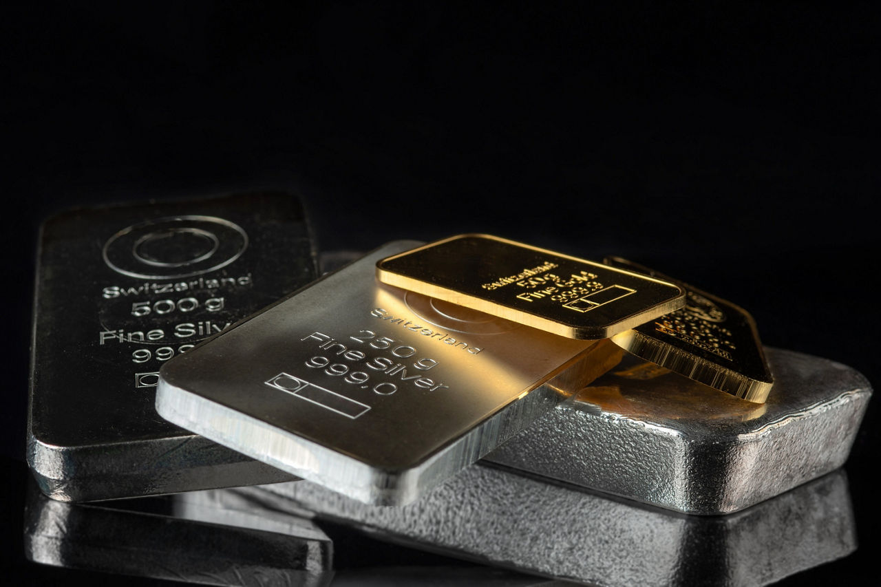 Silber- und Goldbarren liegen unsortiert auf einem kleine Haufen auf einer verspiegelten Oberfläche. Der Hintergrund ist schwarz.