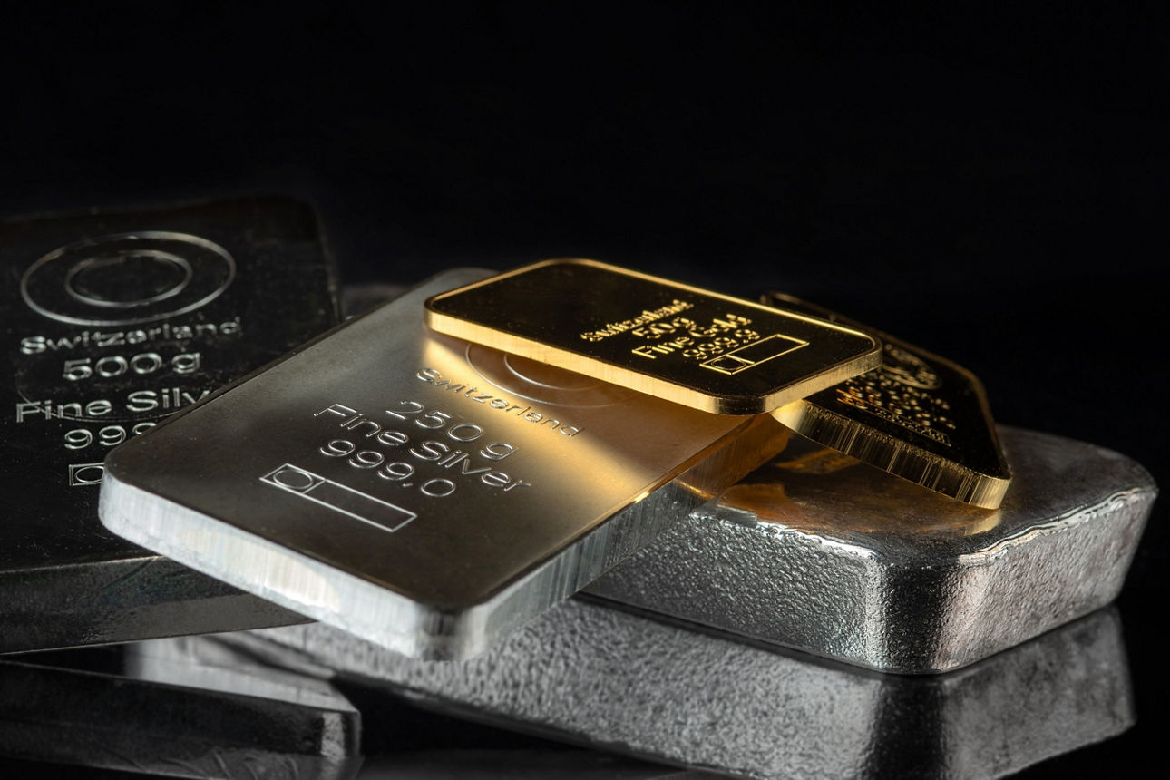 Silber- und Goldbarren liegen unsortiert auf einem kleine Haufen auf einer verspiegelten Oberfläche. Der Hintergrund ist schwarz.