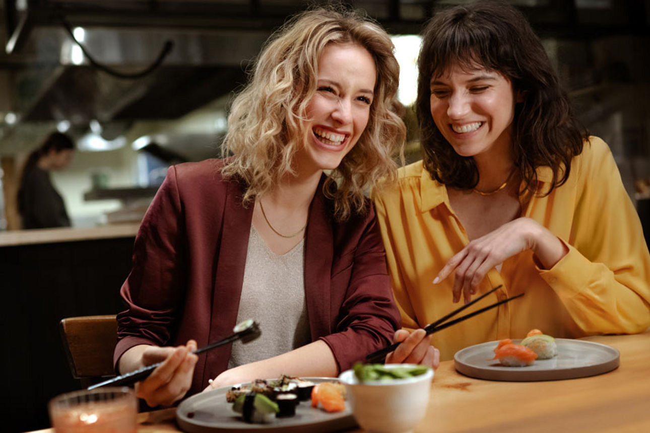 Zwei junge Frauen sitzen lächelnd am Tisch und essen Sushi.