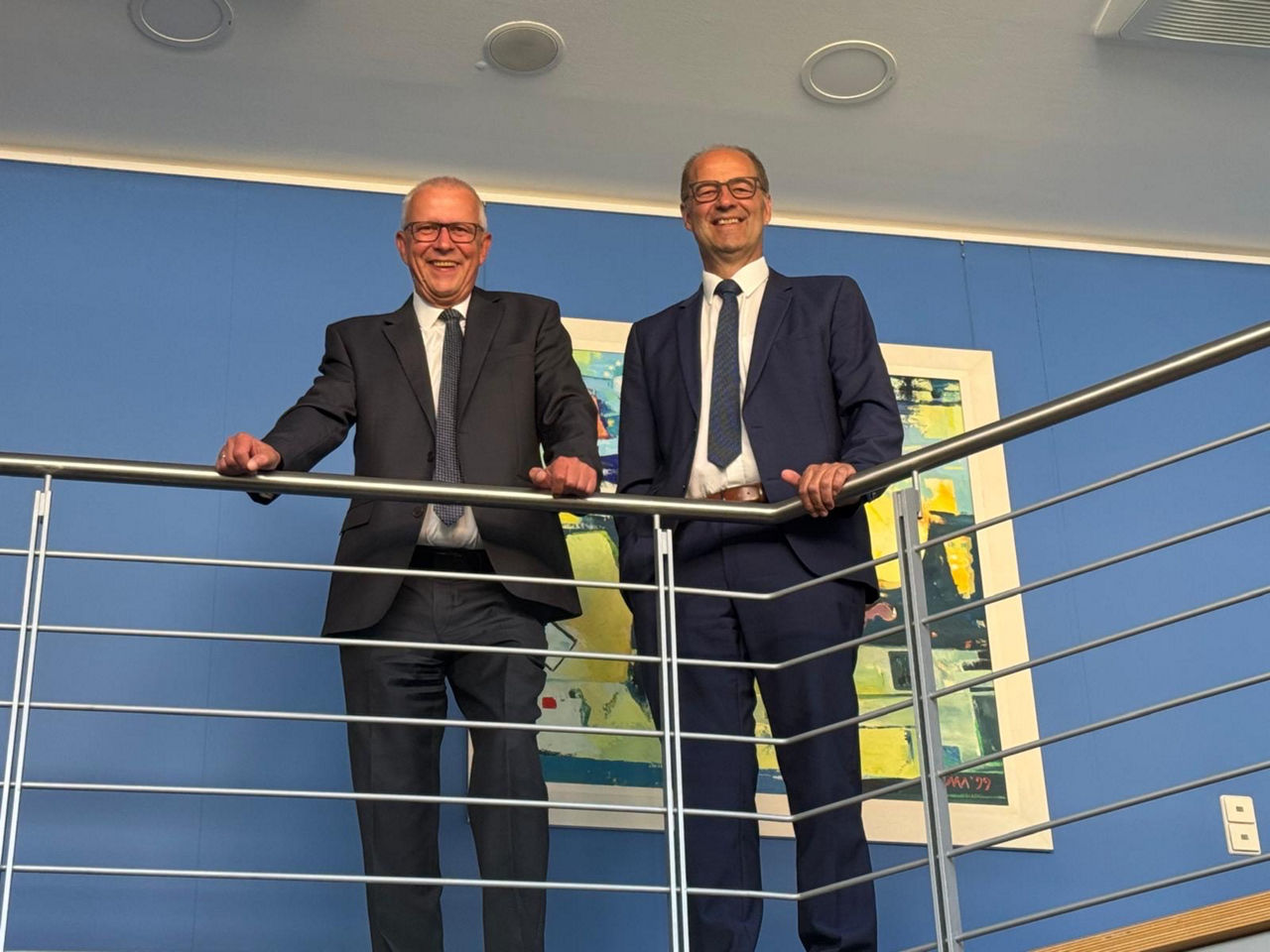 Zwei Männer im Anzug stehen lächelnd an einem Geländer in einem modernen Bürogebäude.