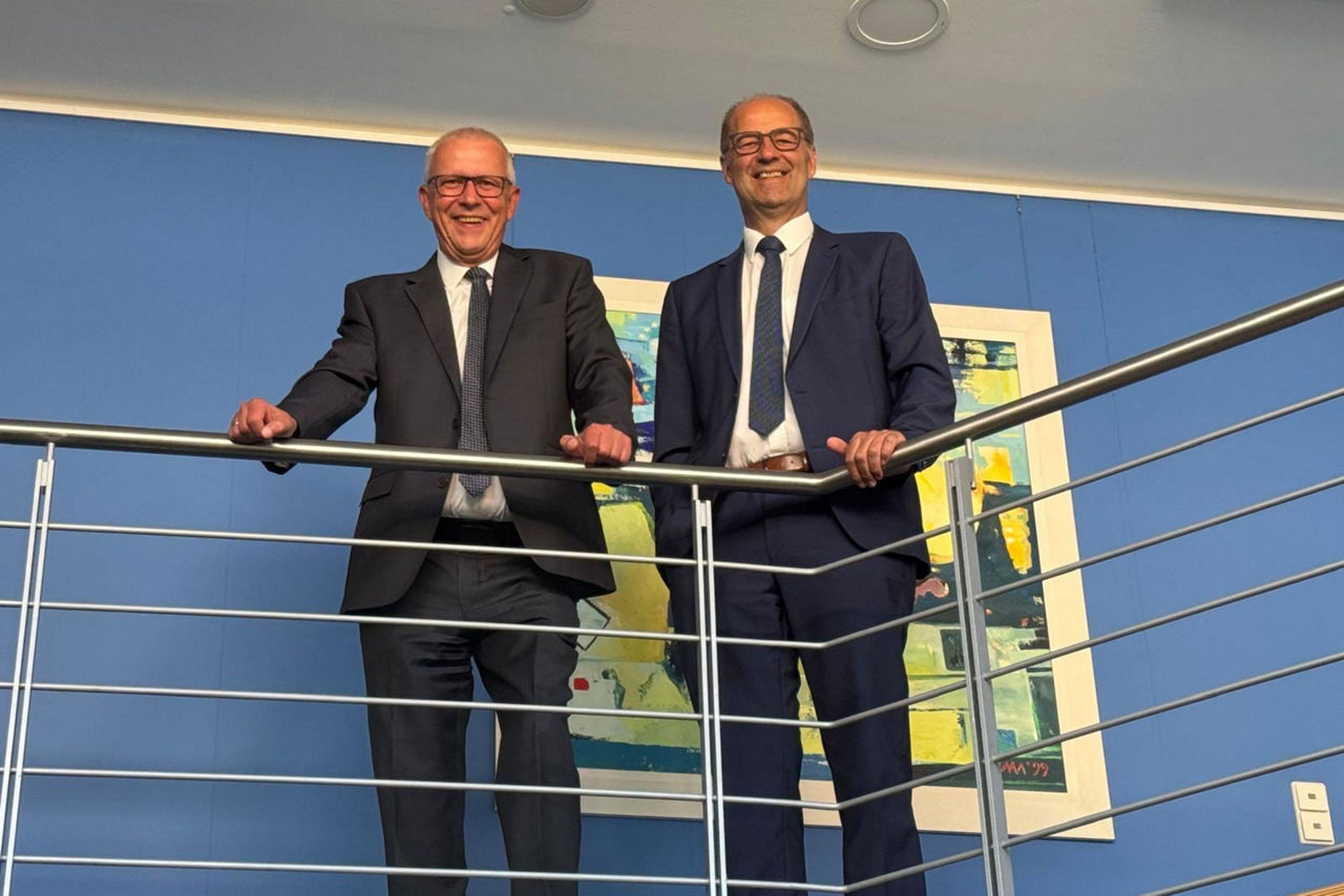 Zwei Männer im Anzug stehen lächelnd an einem Geländer in einem modernen Bürogebäude.