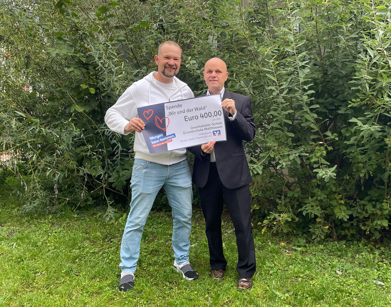 Zwei Erwachsene stehen auf einer Wiese und halten einen Spendenscheck in der Hand.