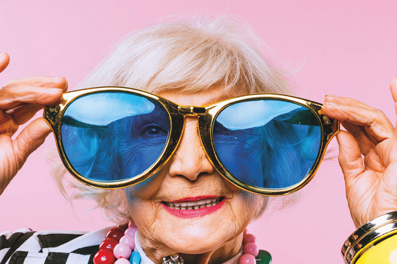 Eine ältere Dame zieht eine übergroße Brille auf. Die Dame trägt bunten, übergroßen Schmuck