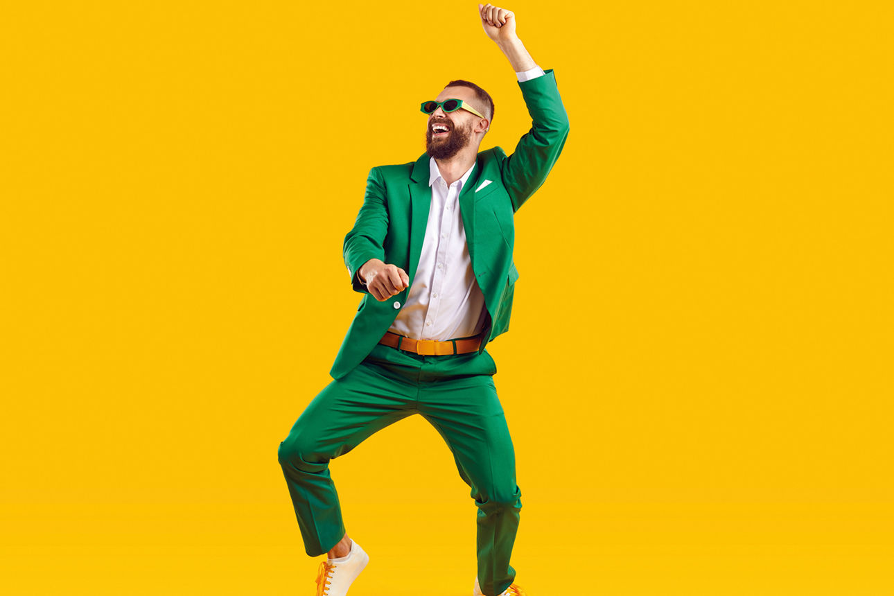 Happy young guy having fun at crazy St Patrick's Day holiday party. Full body shot of funny, cheerful, goofy man wearing green suit and sunglasses dancing isolated on bright yellow colour background