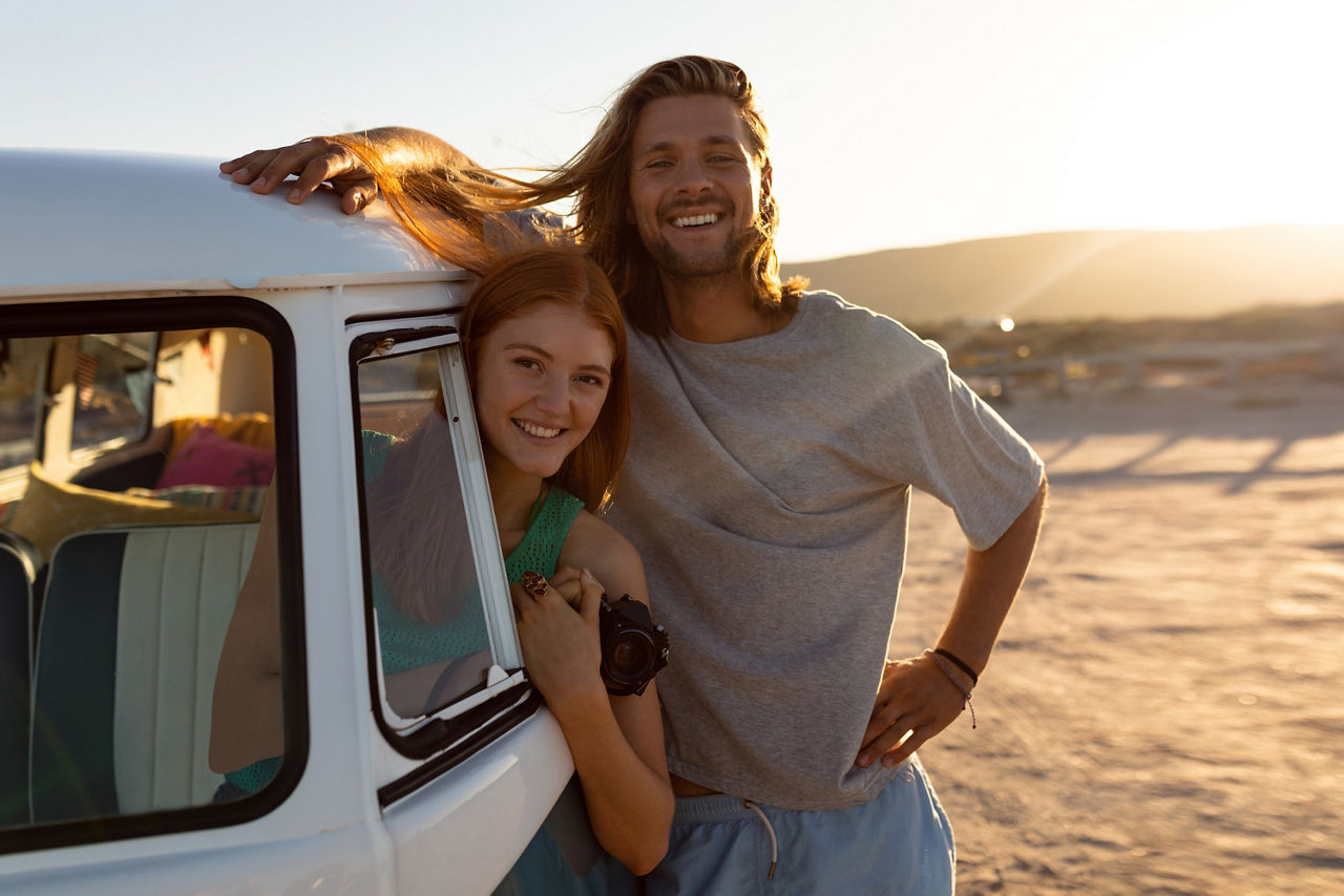 Ein Pärchen parkt mit ihrem Kleinbus am Strand und blickt zufrieden in die Kamera. 
