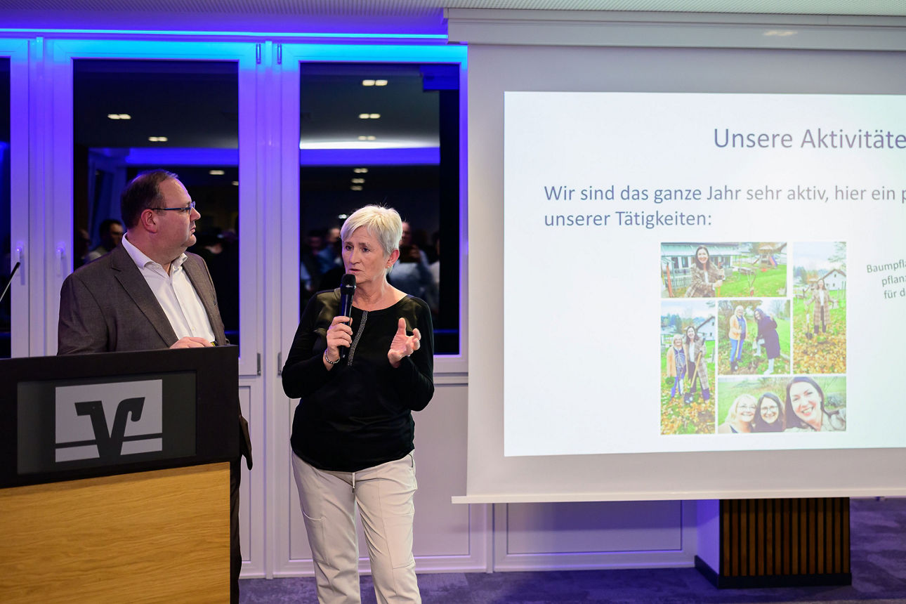 Ein Mann steht beim Rednerpult während eine Frau einen Vortrag hält. Im Hintergrund die Leinwand