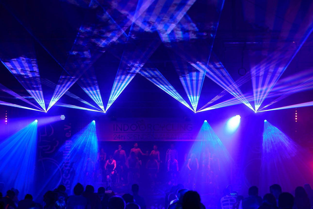 Gruppe von Menschen, die auf einer Bühne in buntem Scheinwerferlicht Indoor-Fahrrad fahren