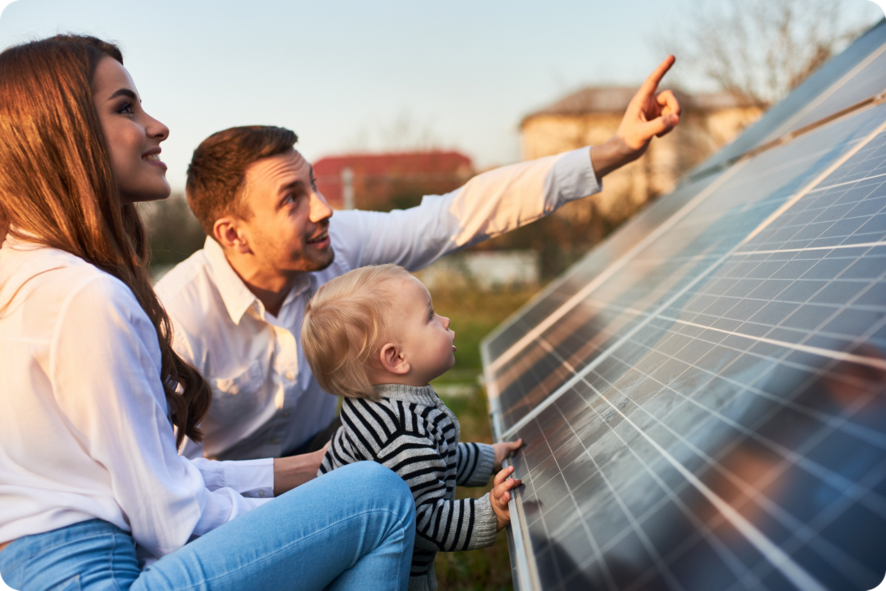 Ein Mann, eine Frau und ein kleines Kind hocken vor einem Solarpanel, der Mann zeigt mit der Hand nach oben