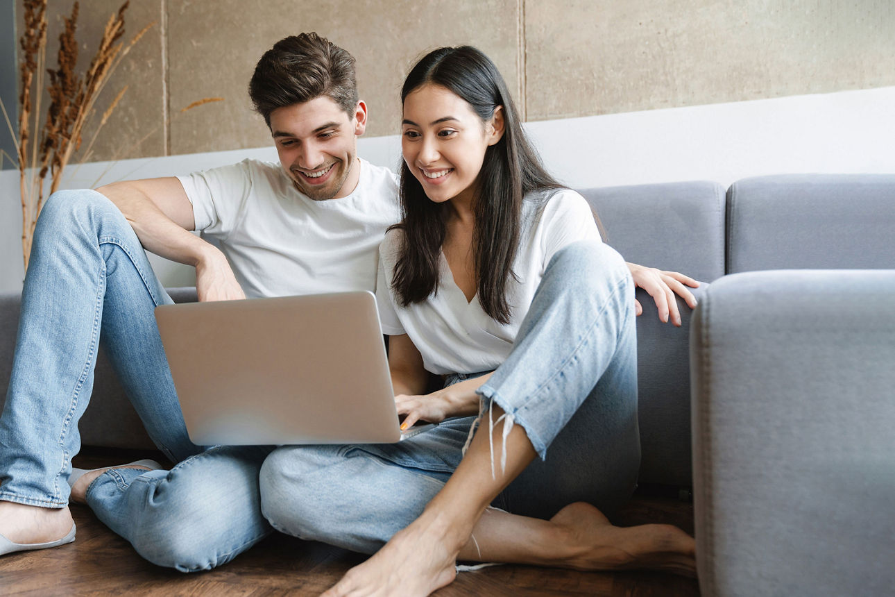 Paar sitzt auf dem Boden vor dem Sofa und nutzt gemeinsam einen Laptop