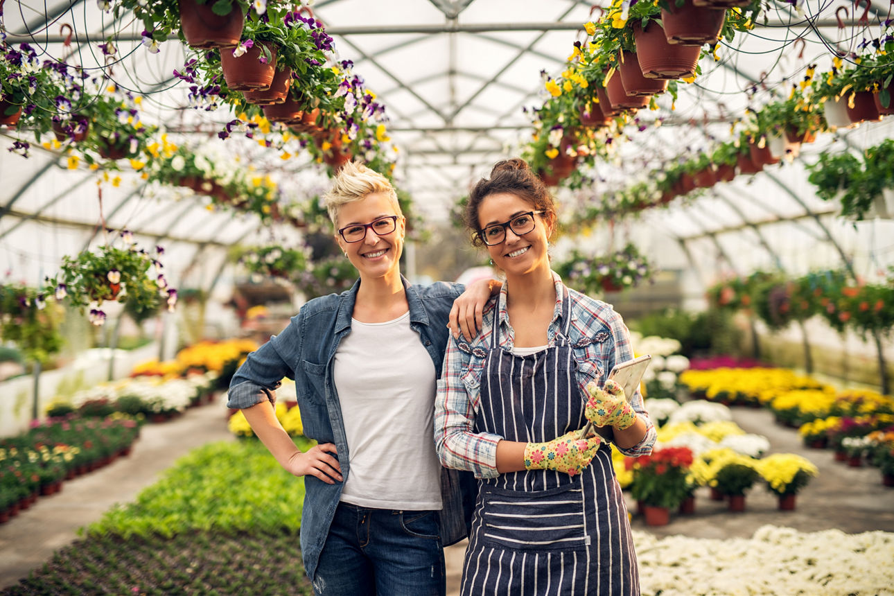 Charming pretty smiling florist woman and female manager posing in a greenhouse full of pots and colourful flowers and looking at the camera.