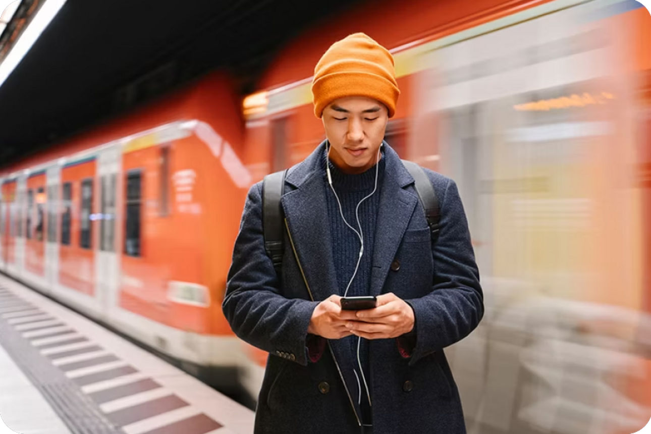 Person wartet mit Kopfhörern an der U‑Bahn und blickt auf ihr Smartphone