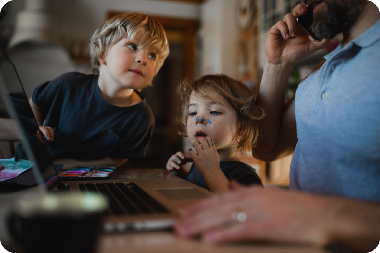Zwei Kinder malen am Laptop, daneben telefoniert eine erwachsene Person.
