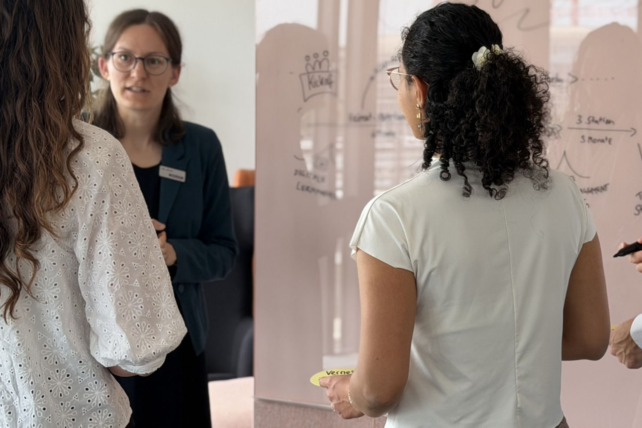 3 junge Menschen stehen vor einem Whiteboard, eine weitere Person präsentiert