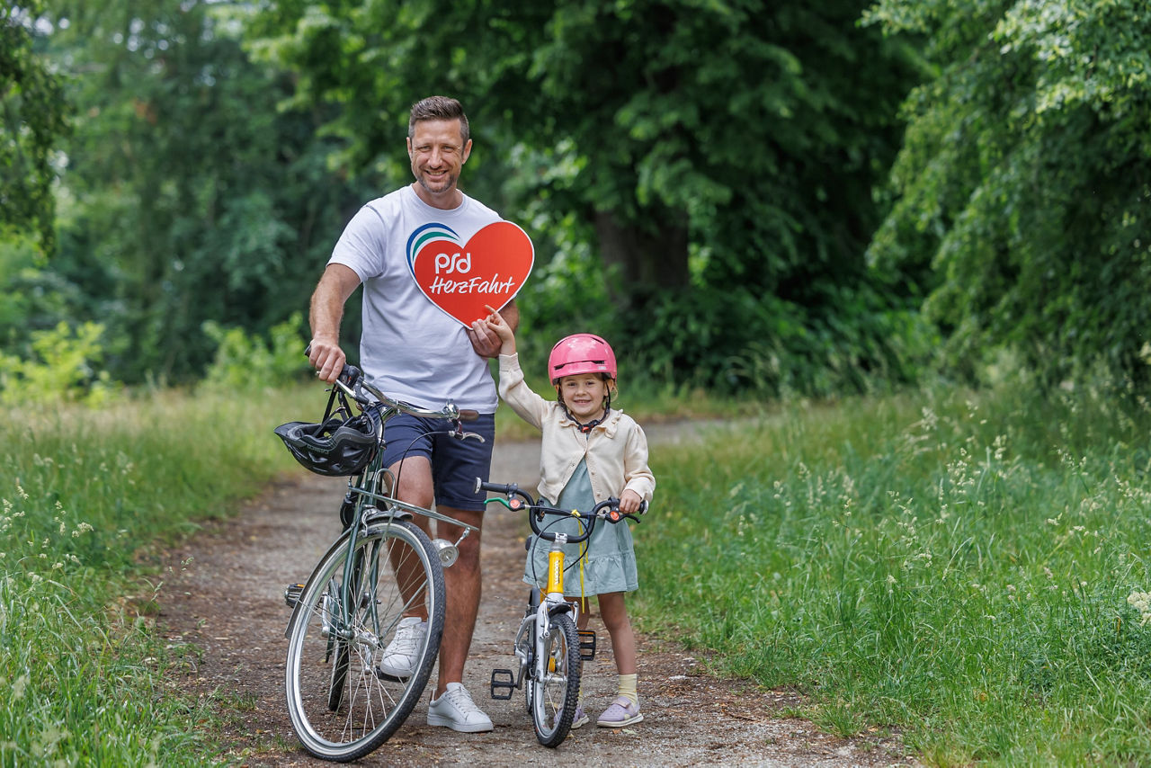 Vater und Tochter mit Fahrrädern halten Logo des PSD Spendenradelns in die Kamera