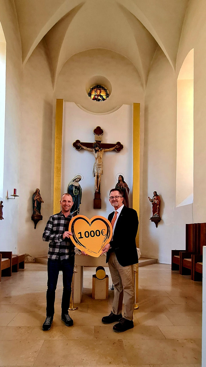 Das Bild zeigt zwei Männer in einer Kirche mit einem herzförmigen Schild mit der Aufschrift "1.000 €".