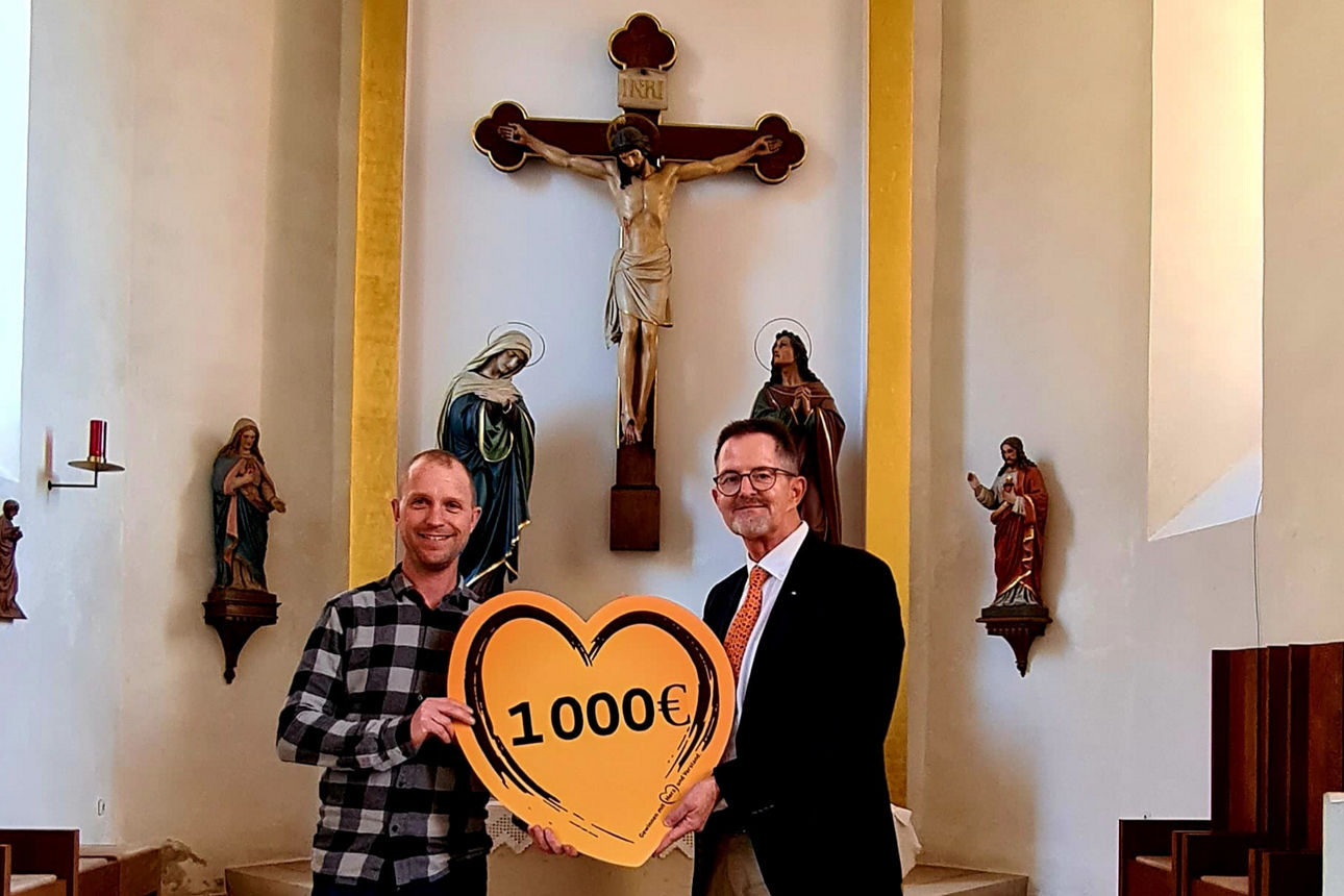 Das Bild zeigt zwei Männer in einer Kirche mit einem herzförmigen Schild mit der Aufschrift "1.000 €".