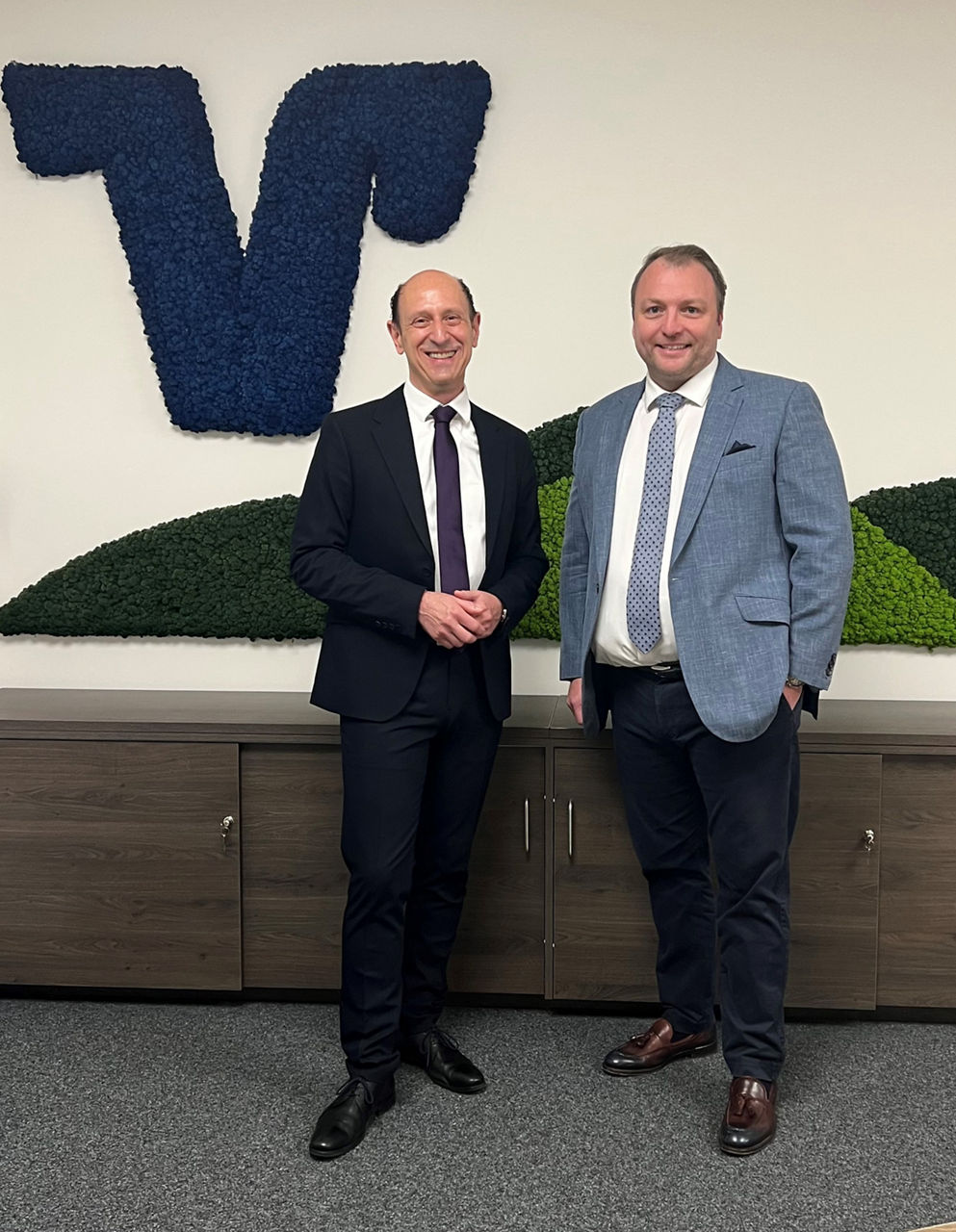 Zwei Männer im Anzug stehen lächelnd in einem modernen Bürogebäude.