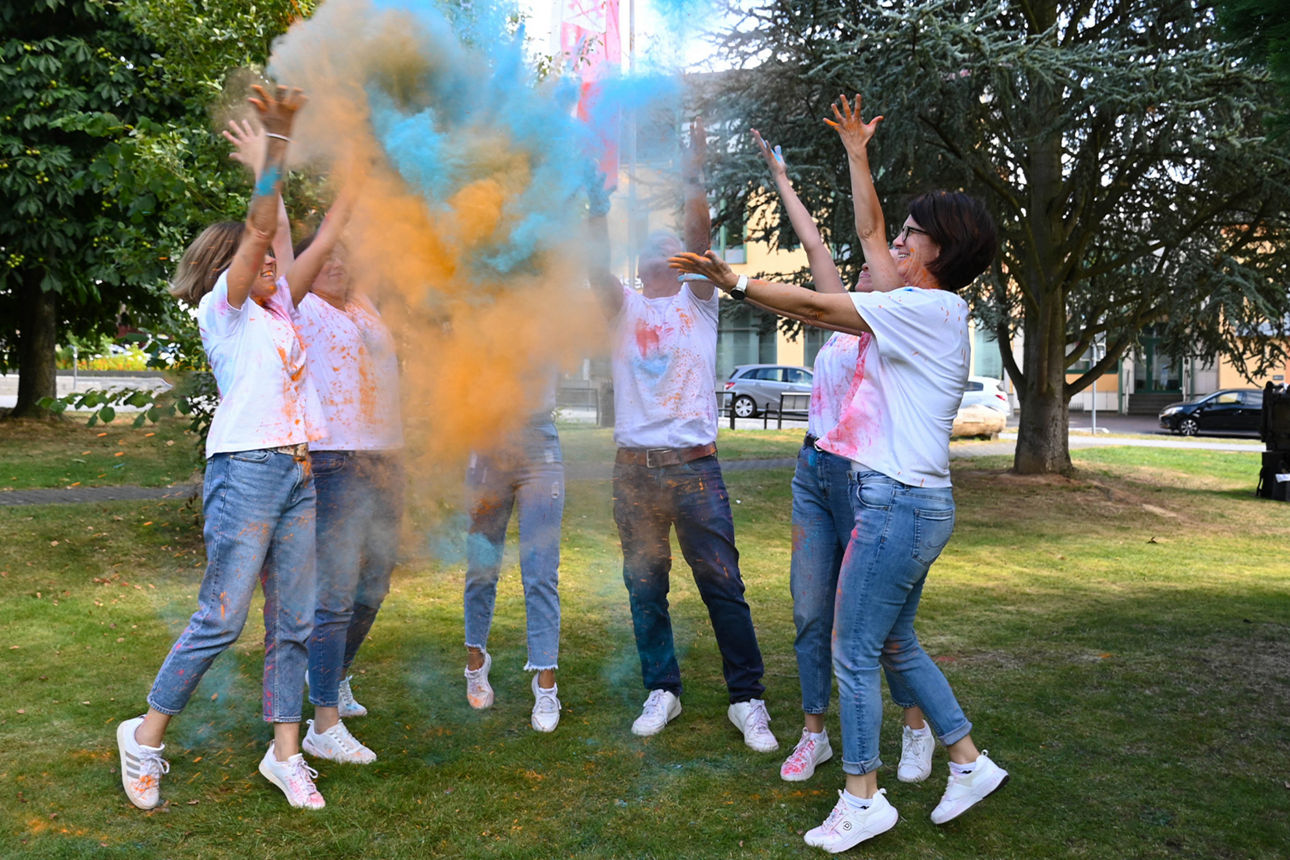 Junge Mitarbeitende der Raiffeisenbank stehen in einem Park und werfen blau-orangefarbenes Holi-Pulver hoch