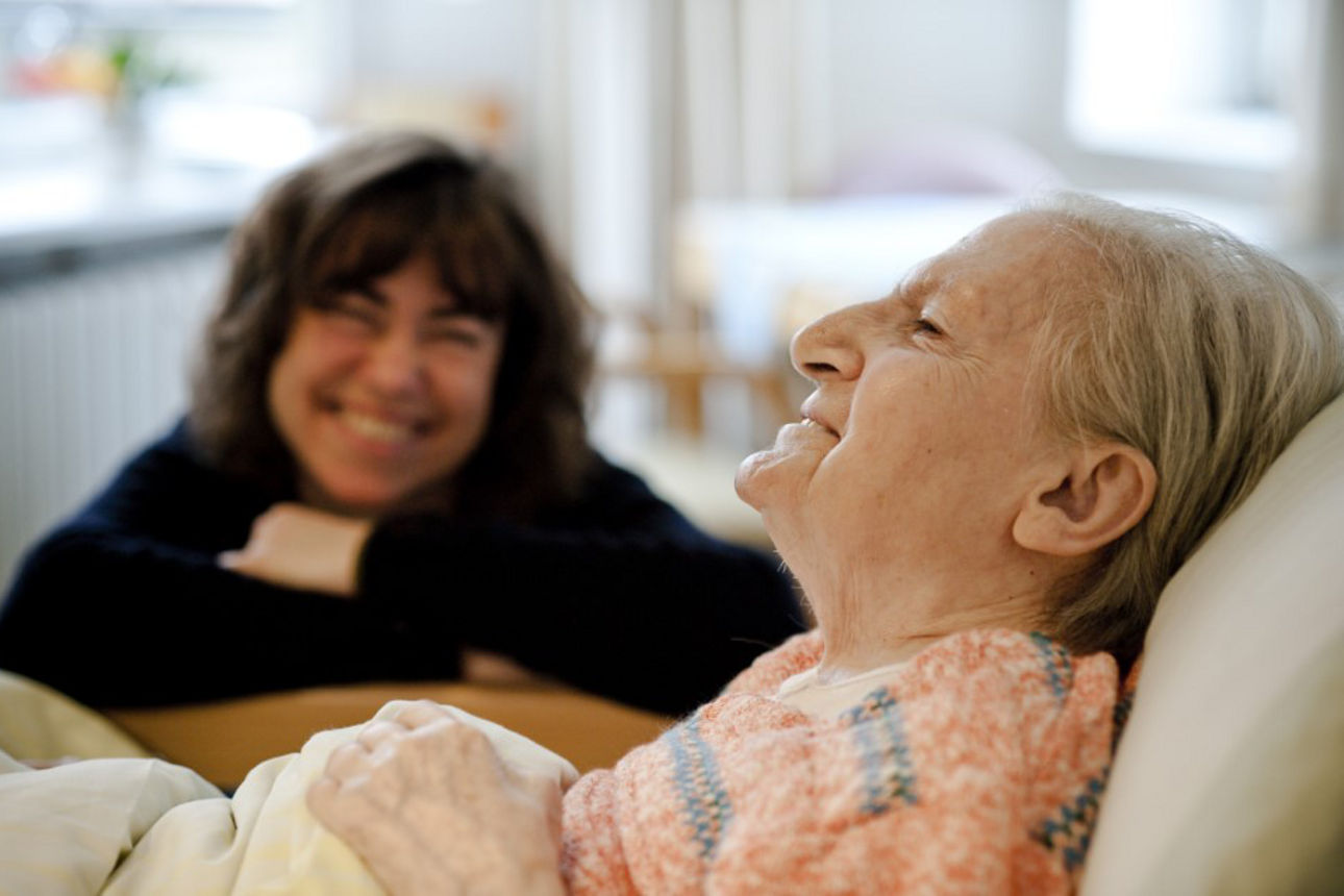 Alte Frau liegt im Bett und lächelt. An der Bettkante hinter ihr sitzt eine junge Frau, die sie anlächelt.