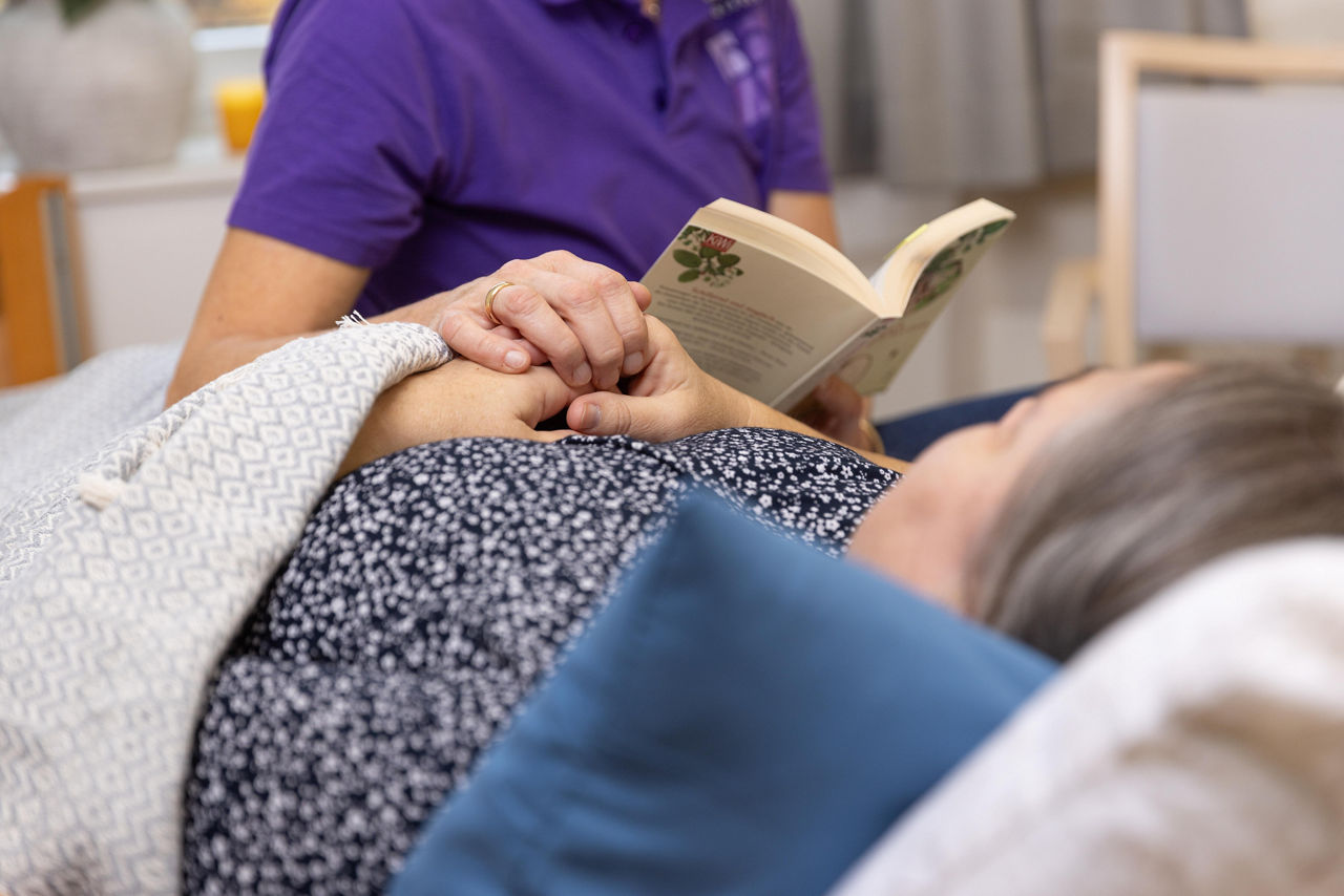 Eine ältere Dame liegt im Bett und bekommt aus einem Buch von einer Pflegerin vorgelesen.
