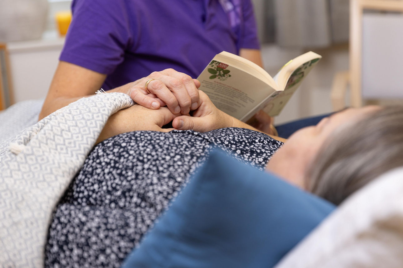 Eine ältere Dame liegt im Bett und bekommt etwas von einer Pflegerin vorgelesen.