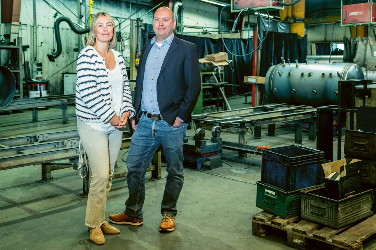 Ein Mann und eine Frau stehen in einer Werkhalle mit Maschinen im Hintergrund und lächeln in die Kamera