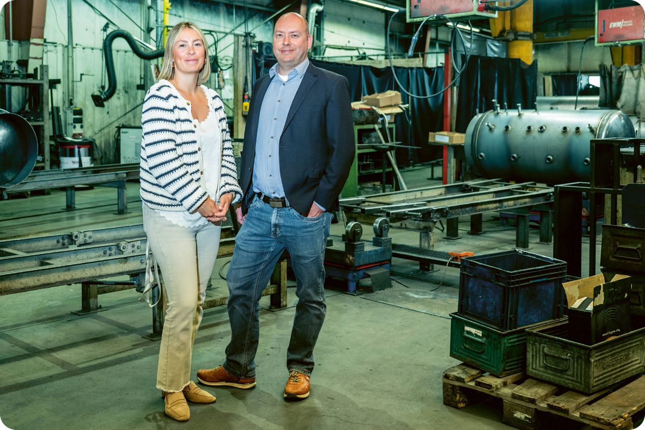 Ein Mann und eine Frau stehen in einer Werkhalle mit Maschinen im Hintergrund und lächeln in die Kamera