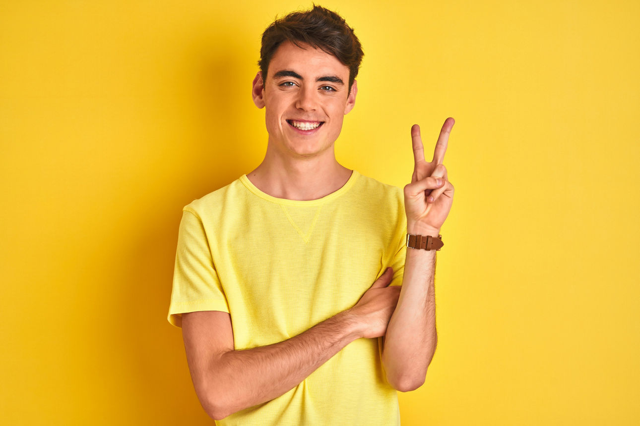 Braunhaariger junger Mann mit gelbem Shirt lächelt und zeigt Peace-Zeichen, Studio Portrait vor gelbem Hintergrund