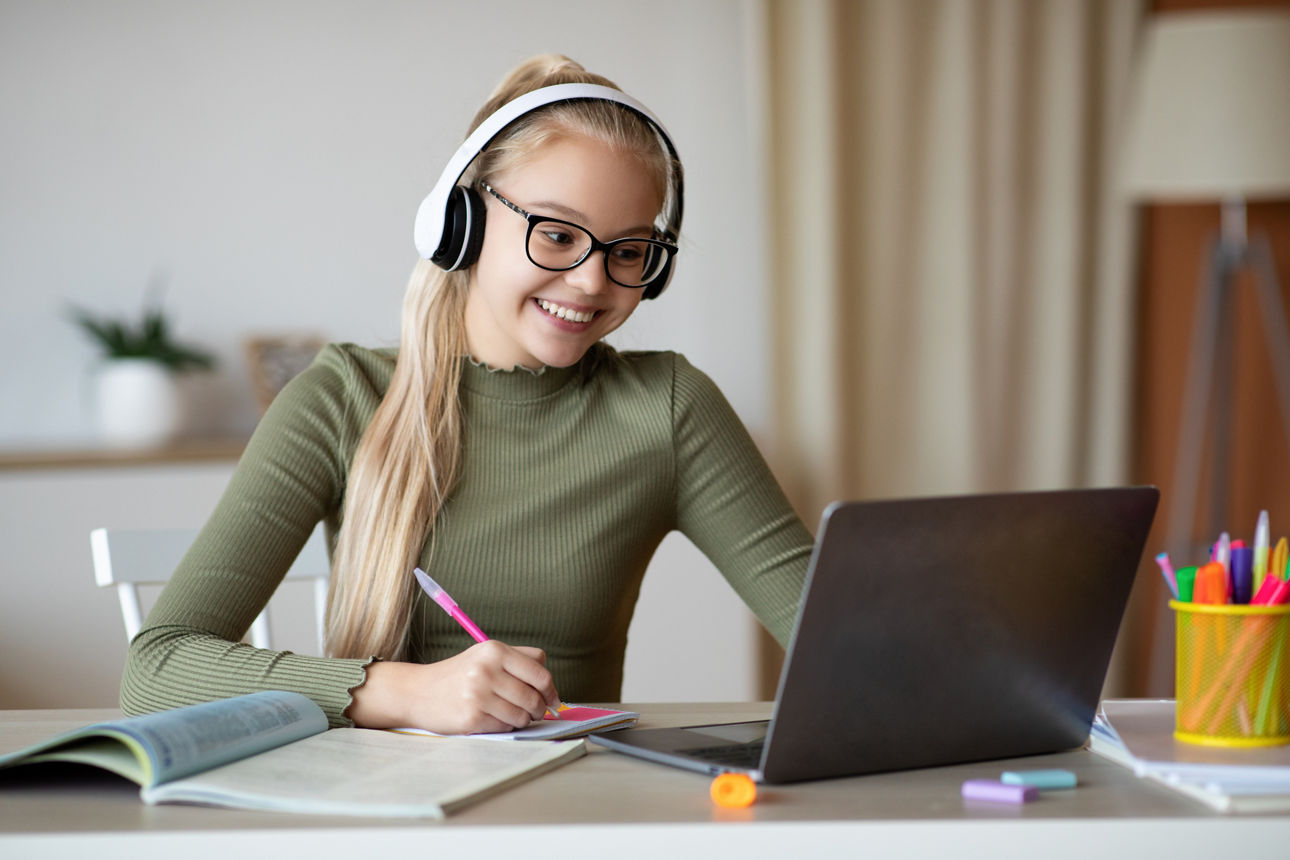 Positive school girl in cute glasses and wireless headset studying from home in front of laptop, looking at computer screen and taking notes, copy space. E-education, online leraning concept