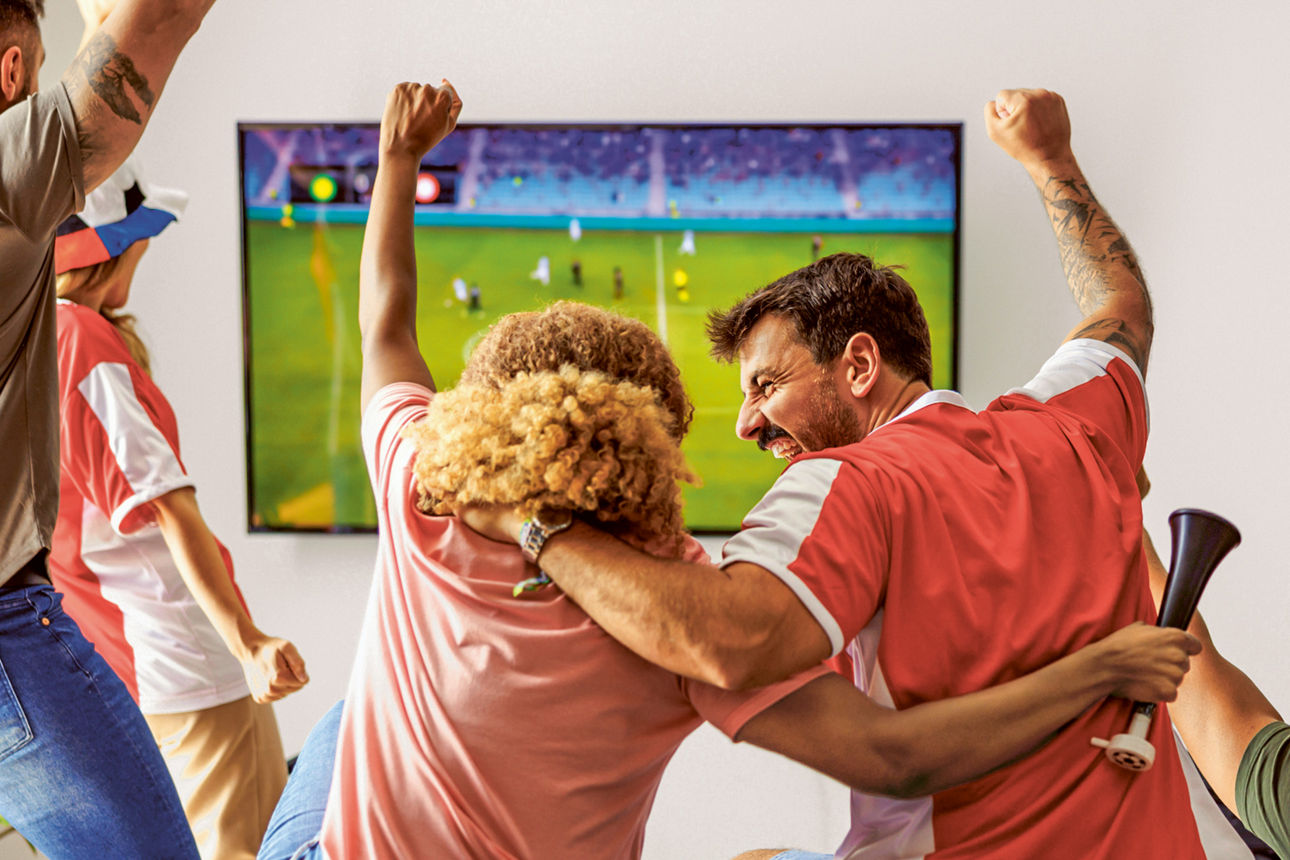 Mehrere Fußballfans jubeln gemeinsam vor einem Fernseher, auf dem ein Spiel läuft, und reißen begeistert die Arme in die Höhe