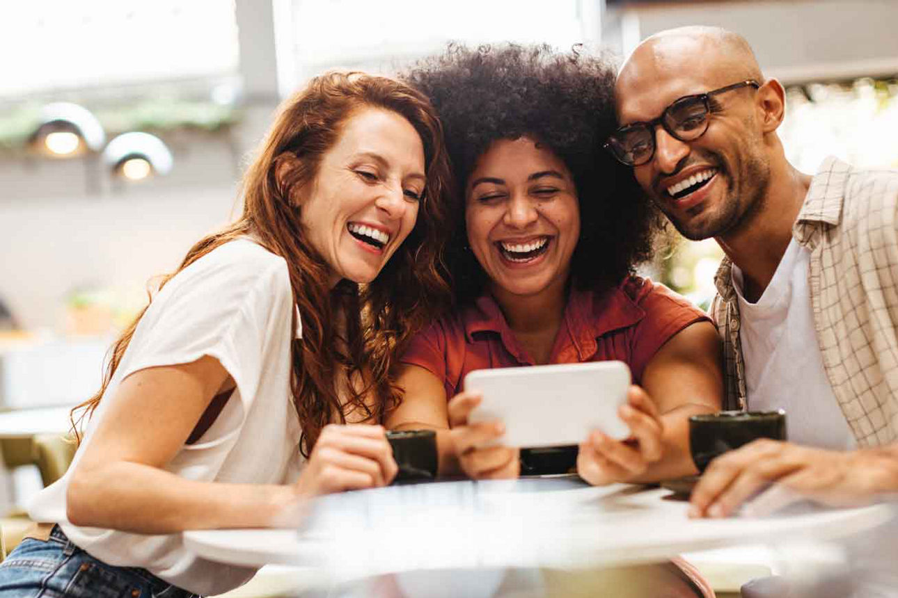 Drei Freunde lachen gemeinsam und schauen auf ein Smartphone am Tisch.