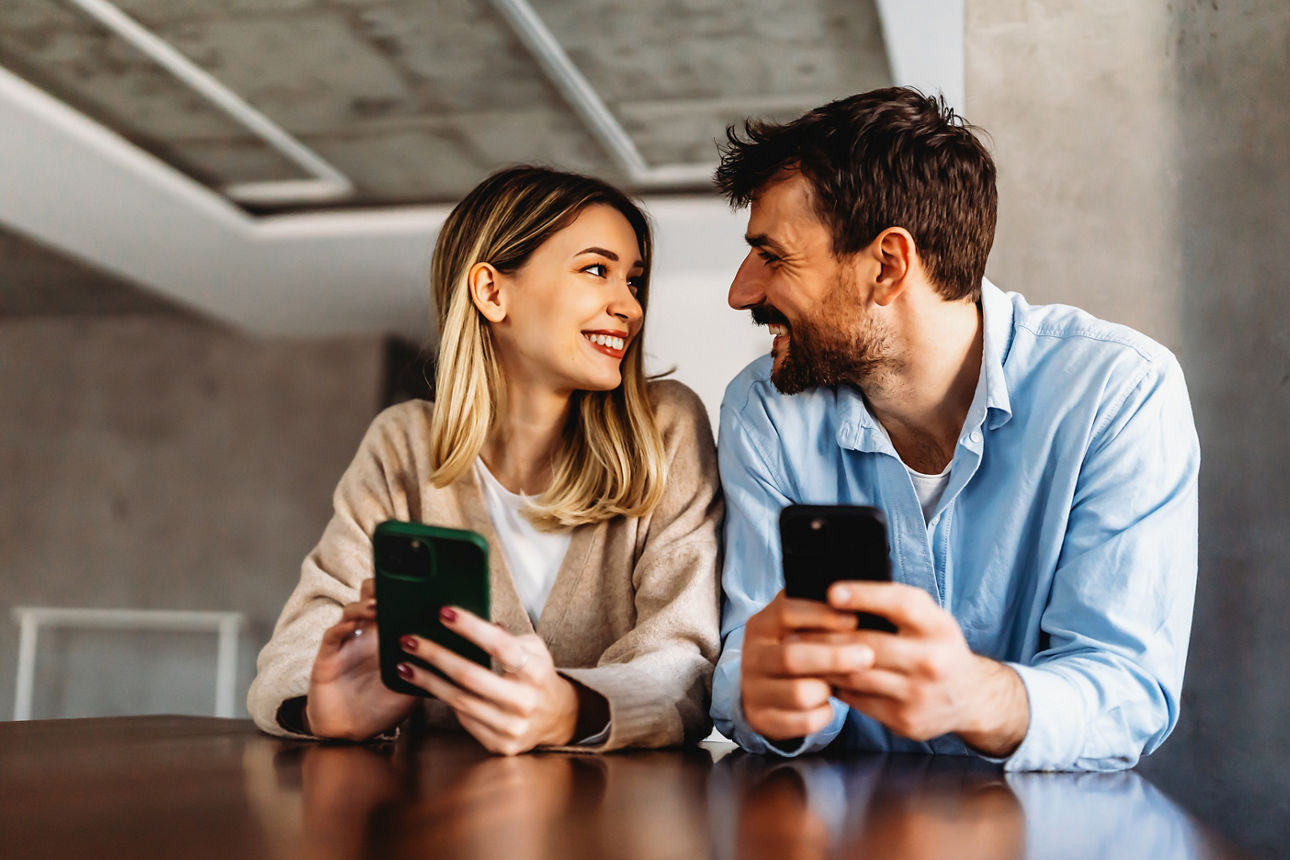 Ein lächelndes junges Paar umarmt sich, während es auf ein Smartphone schaut. Ein multiethnisches Paar betrachtet gemeinsam Social-Media-Inhalte auf dem Smartphone.