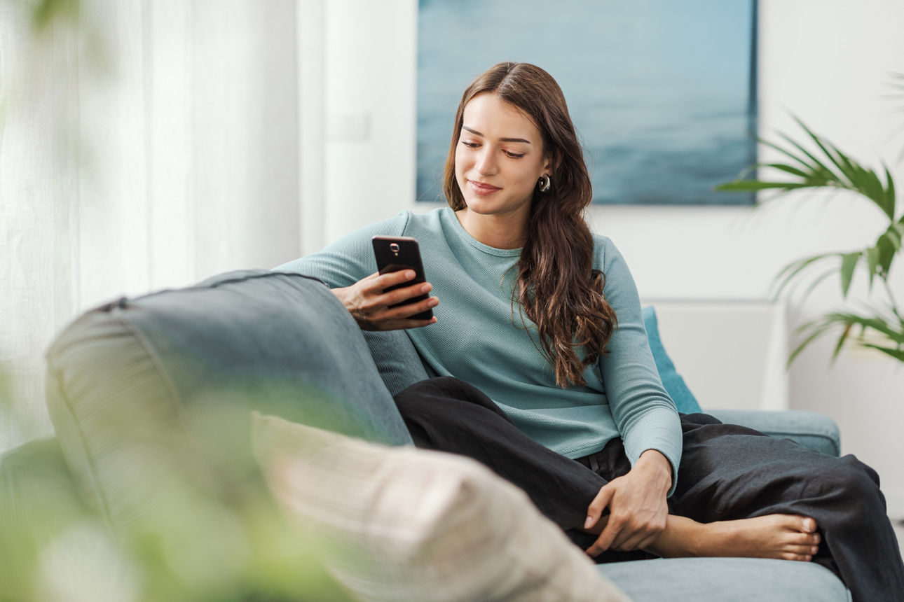 Eine glückliche junge Frau sitzt zu Hause auf dem Sofa und chattet auf ihrem Smartphone; sie ist in sozialen Netzwerken aktiv und nutzt mobile Apps.