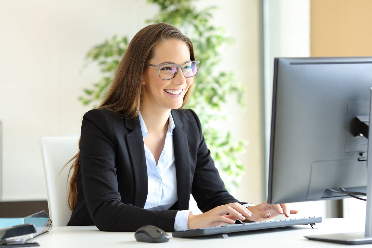 Eine Frau sitzt an einem Schreibtisch in einem modernen Büro und tippt lächelnd auf der Tastatur eines Computers.
