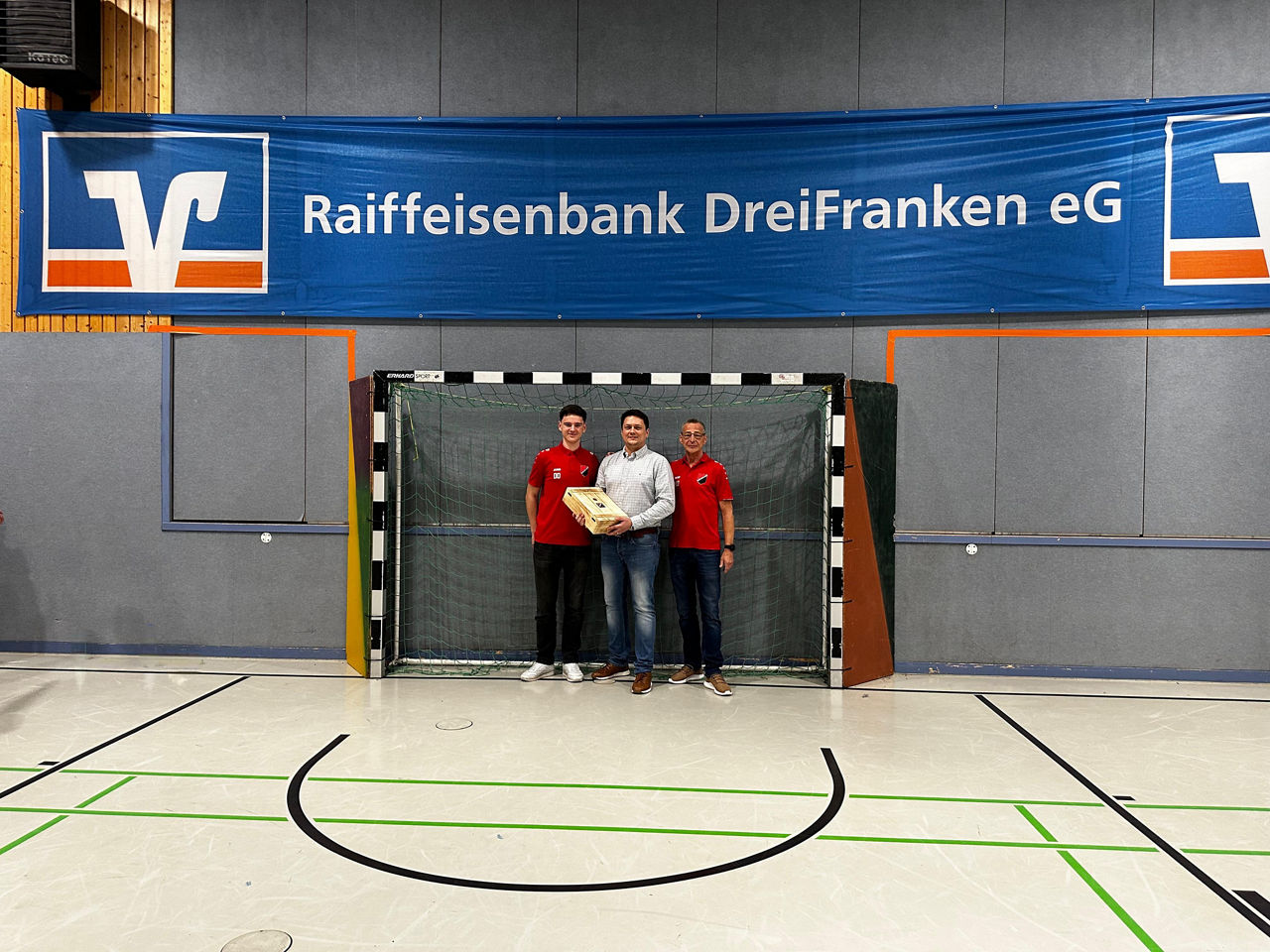Vereinsvorstände und Bankvorstand stehen in einem Fußballtor. Über ihnen hängt ein Banner der Bank.