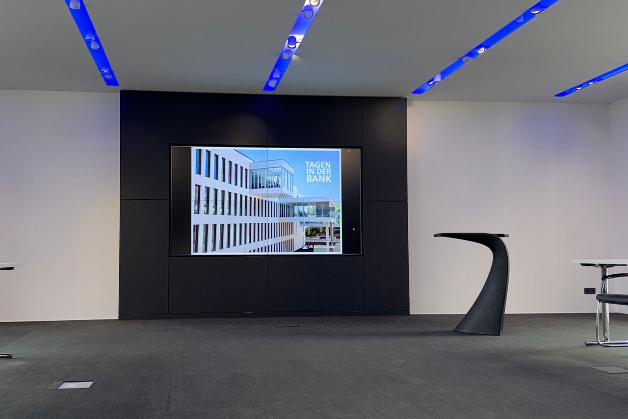 Videowall in einem Konferenzraum, rechts daneben steht ein schwarzes Pult