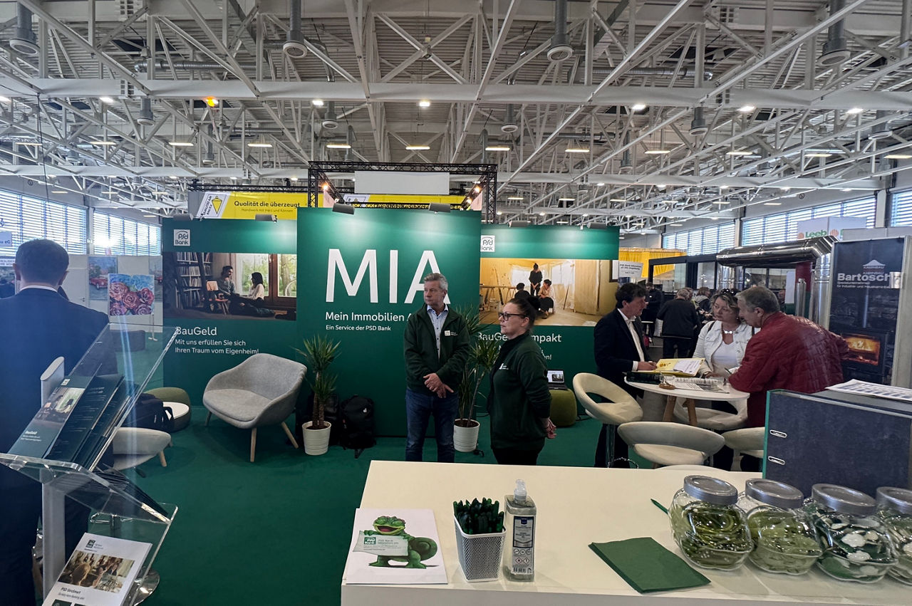 Der Messestand auf den Immobilientagen 2025. In der Mitte steht ein Mitarbeiter und eine Mitarbeiterin, am Rand sieht man Berater im Gespräch mit Kunden.