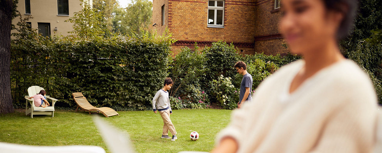 Zwei Kinder spielen auf einer Wiese mit einem Ball. Davor sitzt eine Frau mit ihrem Laptop. Sie befinden sich in einem Garten.