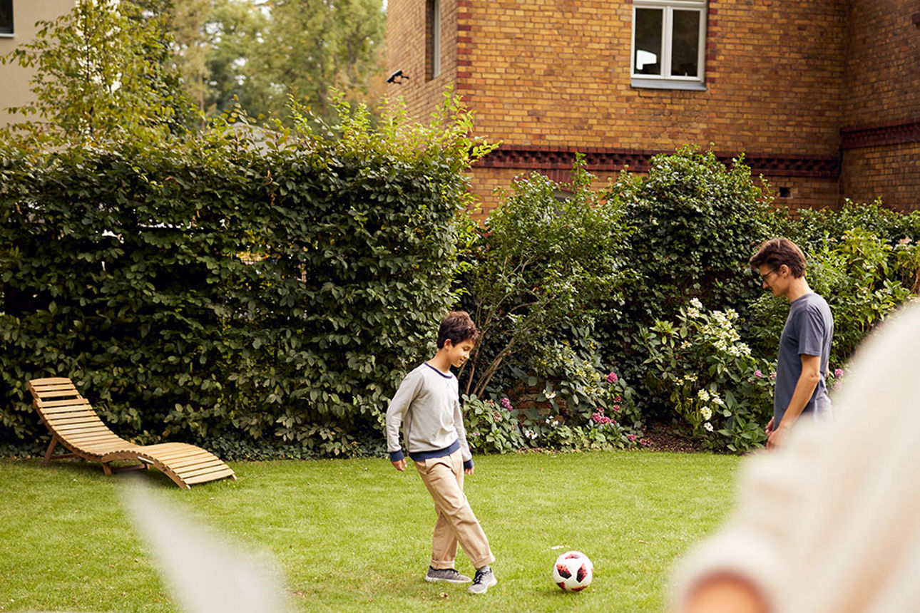 Zwei Kinder spielen auf einer Wiese mit einem Ball. Davor sitzt eine Frau mit ihrem Laptop. Sie befinden sich in einem Garten.