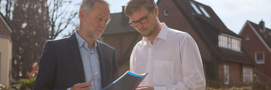 Zwei Männer im Vordergrund eines Hauses die gemeinsam auf einen Plan schauen