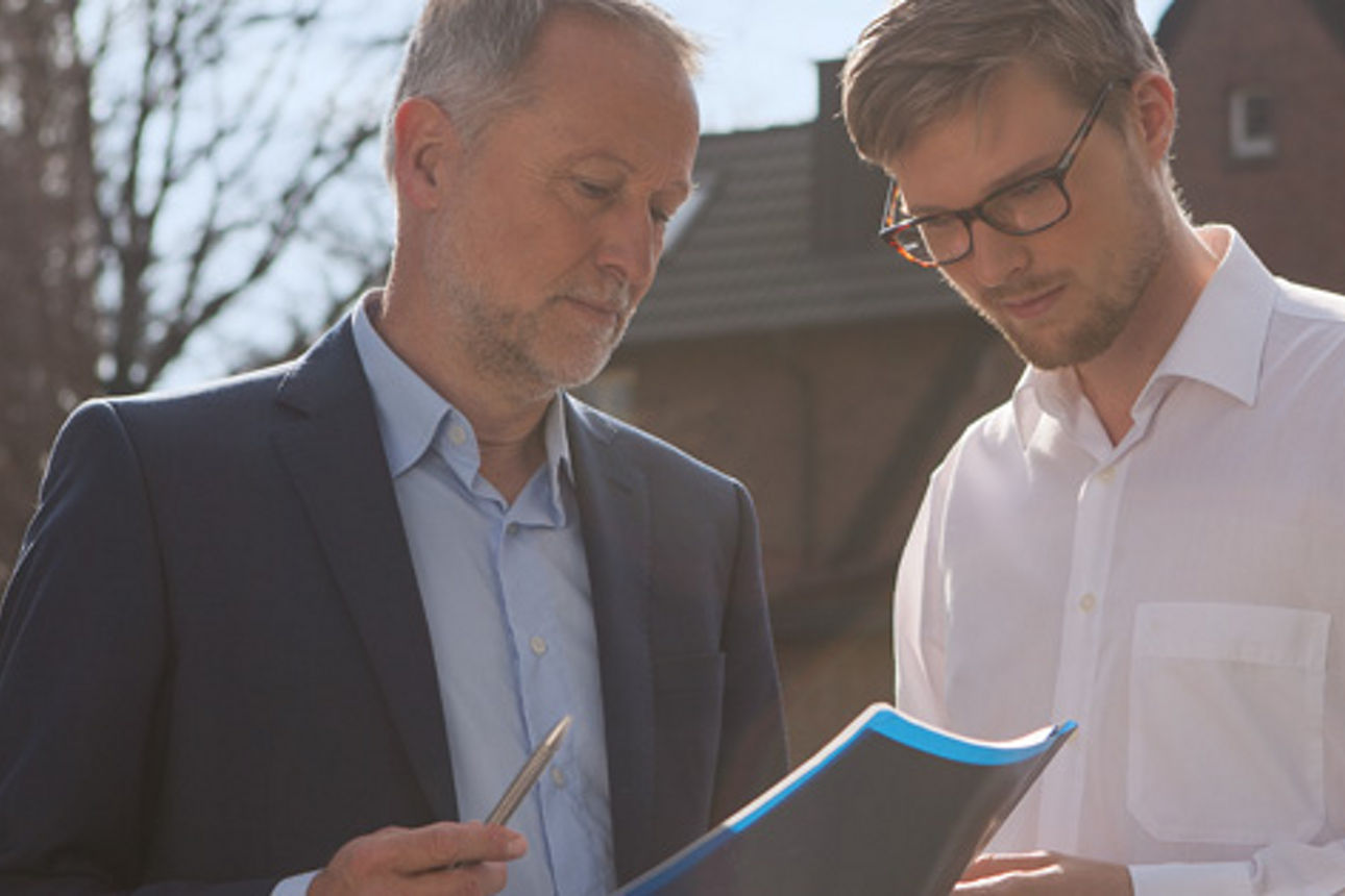 Zwei Männer im Vordergrund eines Hauses die gemeinsam auf einen Plan schauen