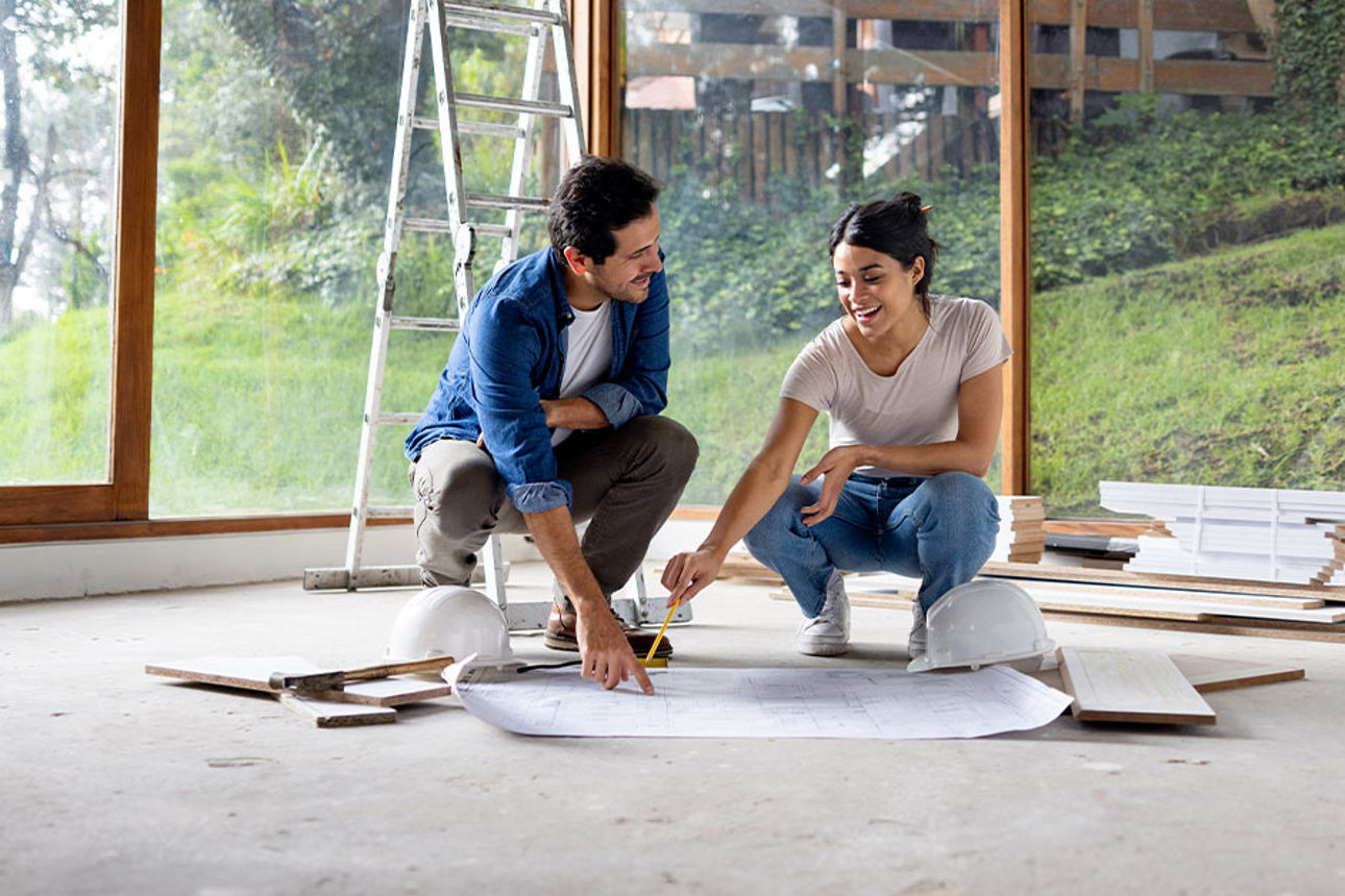 Ein Paar sitz auf dem Boden eines unfertigen Raums und schauen auf Baupläne, umgeben von Baumaterialien und einer Leiter