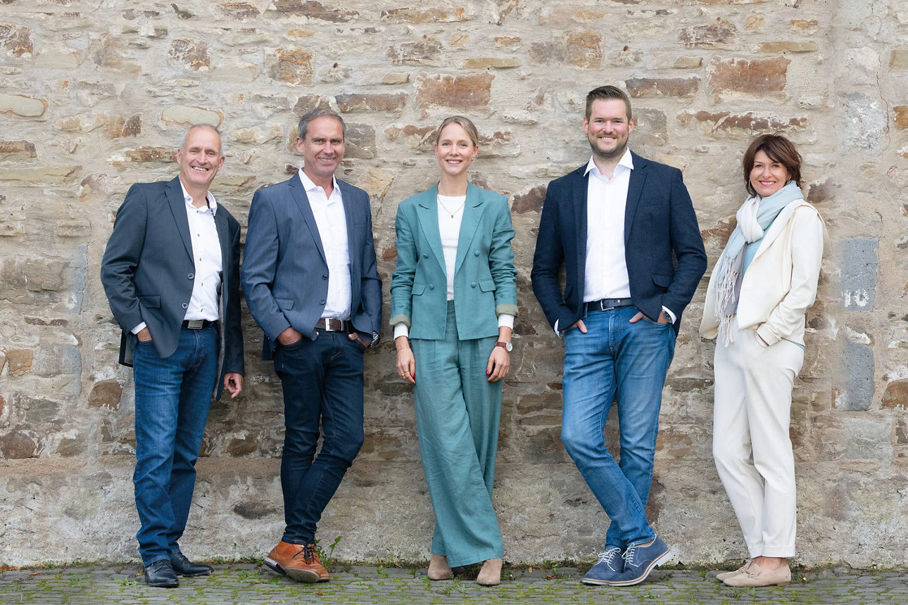 Drei Männer und zwei Frauen stehen vor einer Hauswand und schauen in die Kamera