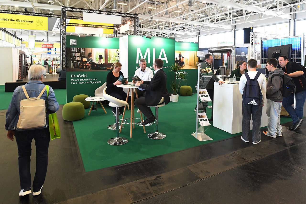 Der Messestand auf den Immobilientagen 2025. In der Mitte ist ein Berater im Kundengespräch mit einem Paar.
