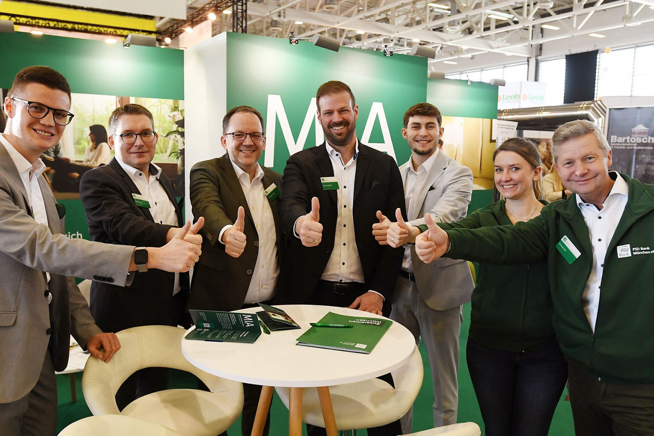 Das Team der PSD Bank München auf den Immobilientagen 2025. Die Mitarbeiter*innen stehen im Halbkreis um einen Tisch und strecken den Daumen in die Kamera